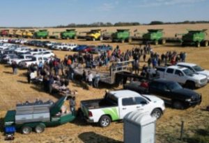People gathered in a field for a Foodgrains harvest. There is a row of green and yellow combines in the back of the photo.