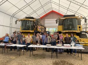 A group of people gathered in front of two yellow combines. Everything is in a tarp quonset.