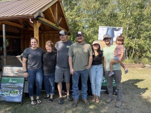 A group of people ranging in age from a toddler up stand for a photo. There are trees and a wooden structure behind them.