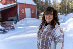 A woman stands in a snow-covered yard. A red building and trees are in the background.