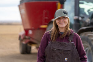 A woman in a hoodie and overalls stands in front of a tractor.