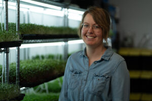 A woman stands in front of an indoor greenhouse, filled with trays and trays of sprouts