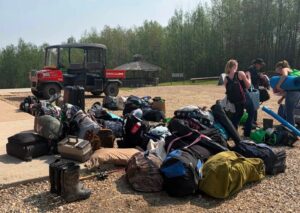 A pile of luggage, boots and other gear with a UTV in the background. Three youth pick up their belongings on the one side