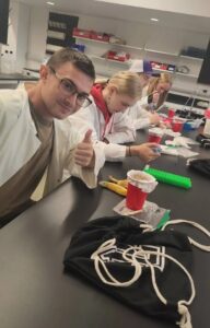 Three youth in lab coats perform experiments while the fourth gives a thumbs up