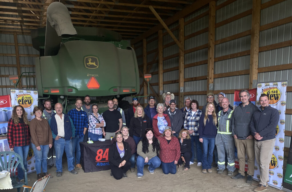 A group of people gathered in a shop for a photo. There is a green combine in the background