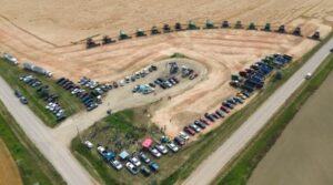 An aerial view of a Canadian Foodgrains harvest event. There are several combines, parked vehicles and people in the shot